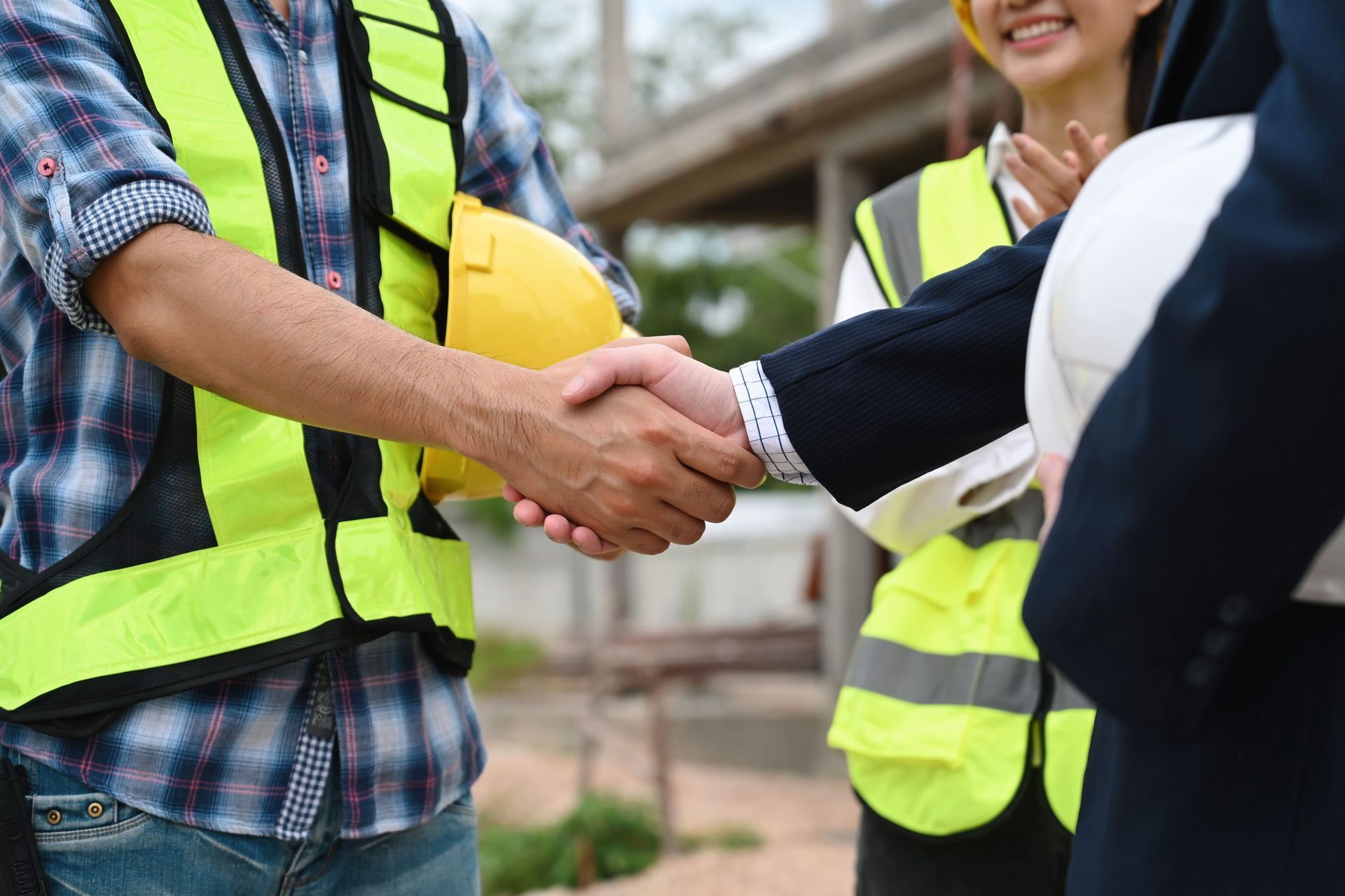 Construction workers shaking hands, signifying agreement. A person wears a safety vest and holds a yellow hard hat. Other workers in the background.