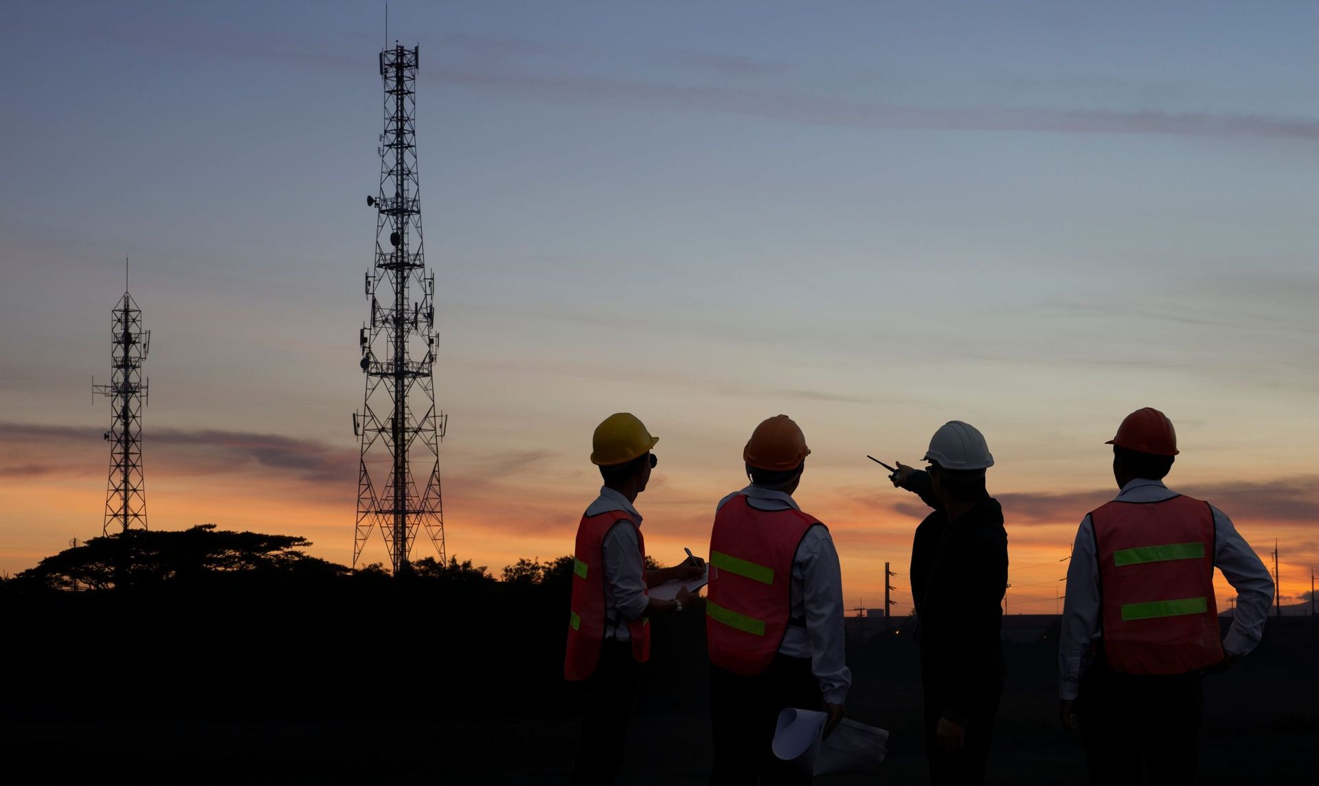 Silhouette of engineers in safety vests and hard hats, inspecting cell towers at sunset.