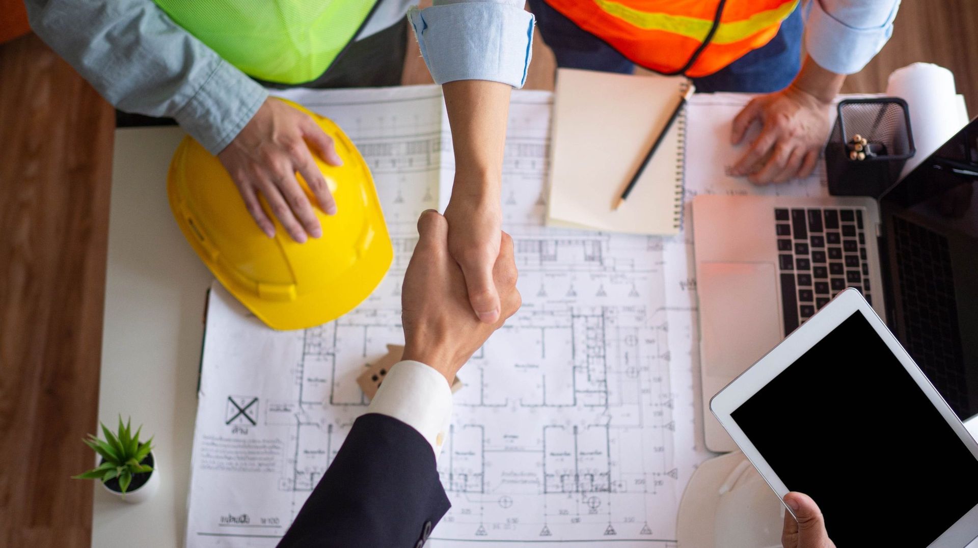Two people shaking hands over architectural plans on a desk, one wearing a suit and the other in construction attire.