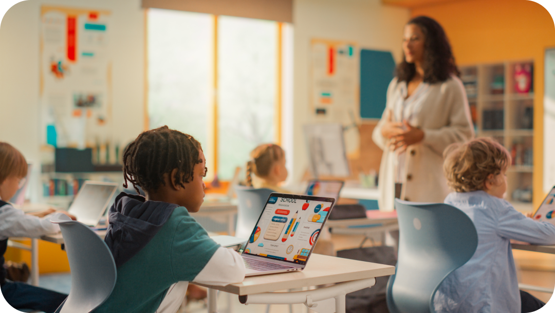 Students in a classroom using laptops while the teacher stands and addresses them.