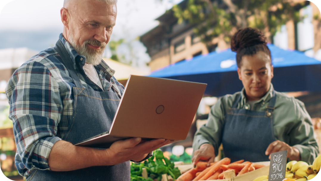 Man with laptop at a market, woman in apron arranging carrots, outdoor setting.