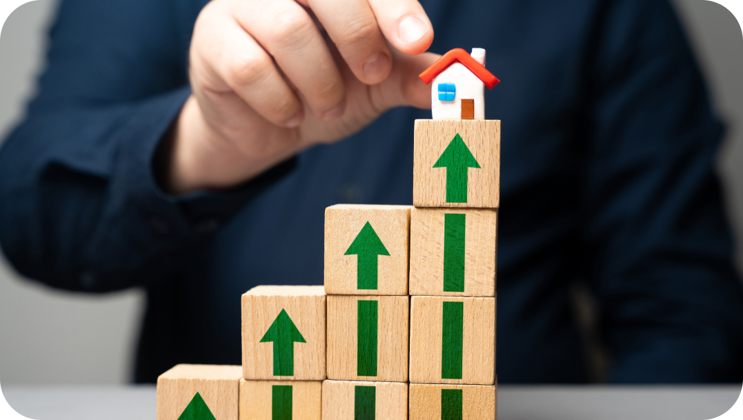 Hand placing a small house on a stack of blocks with upward arrows, symbolizing growth and real estate.