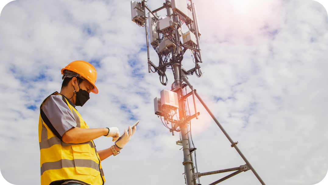 Worker in safety gear inspecting a cell tower, with a cloudy sky in the background.