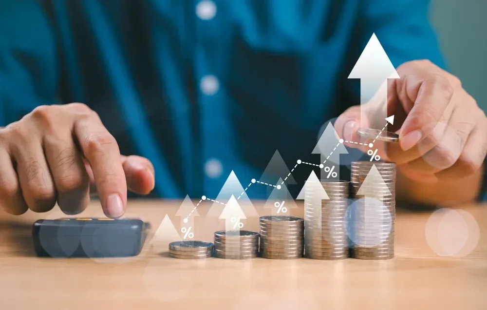 Person calculating, with growing stacks of coins and a rising stock chart.