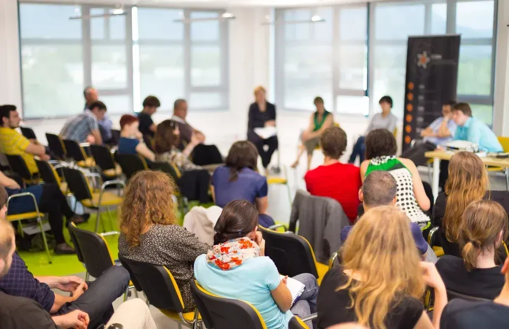 Audience at a panel discussion in a light-filled room; speakers in front; attendees in chairs, listening.