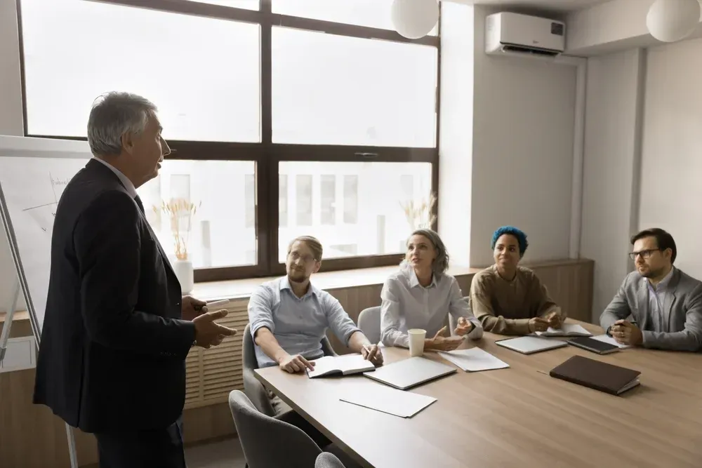 A businessman in suit giving a presentation to a diverse group seated at a conference table in a well-lit office.
