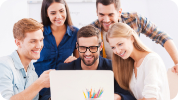 Group of diverse people smiling, gathered around a laptop, appearing to collaborate and share ideas in a bright setting.
