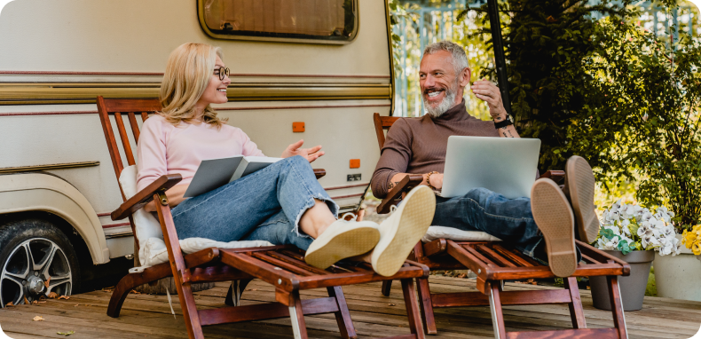 Couple relaxing in chairs, working on laptops next to an RV. Outdoors with trees. Smiling.
