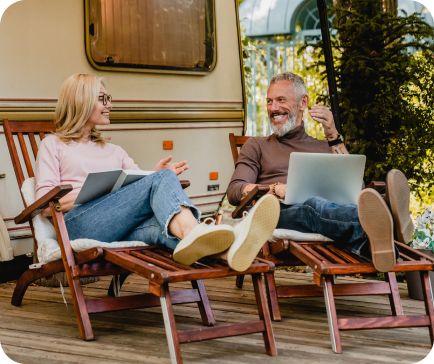 Couple relaxing on lounge chairs by a camper, woman holding a book, man with laptop, smiling. Couple relaxing on lounge chairs by a camper, woman holding a book, man with laptop, smiling.