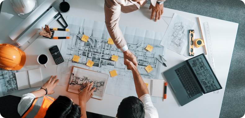 People shaking hands over blueprints and a laptop on a table.