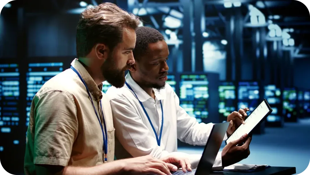 Two IT technicians in data center, looking at a tablet and laptop, analyzing data.