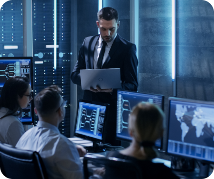 Group of people in a server room, looking at computer monitors. A man in a suit presents with a laptop.