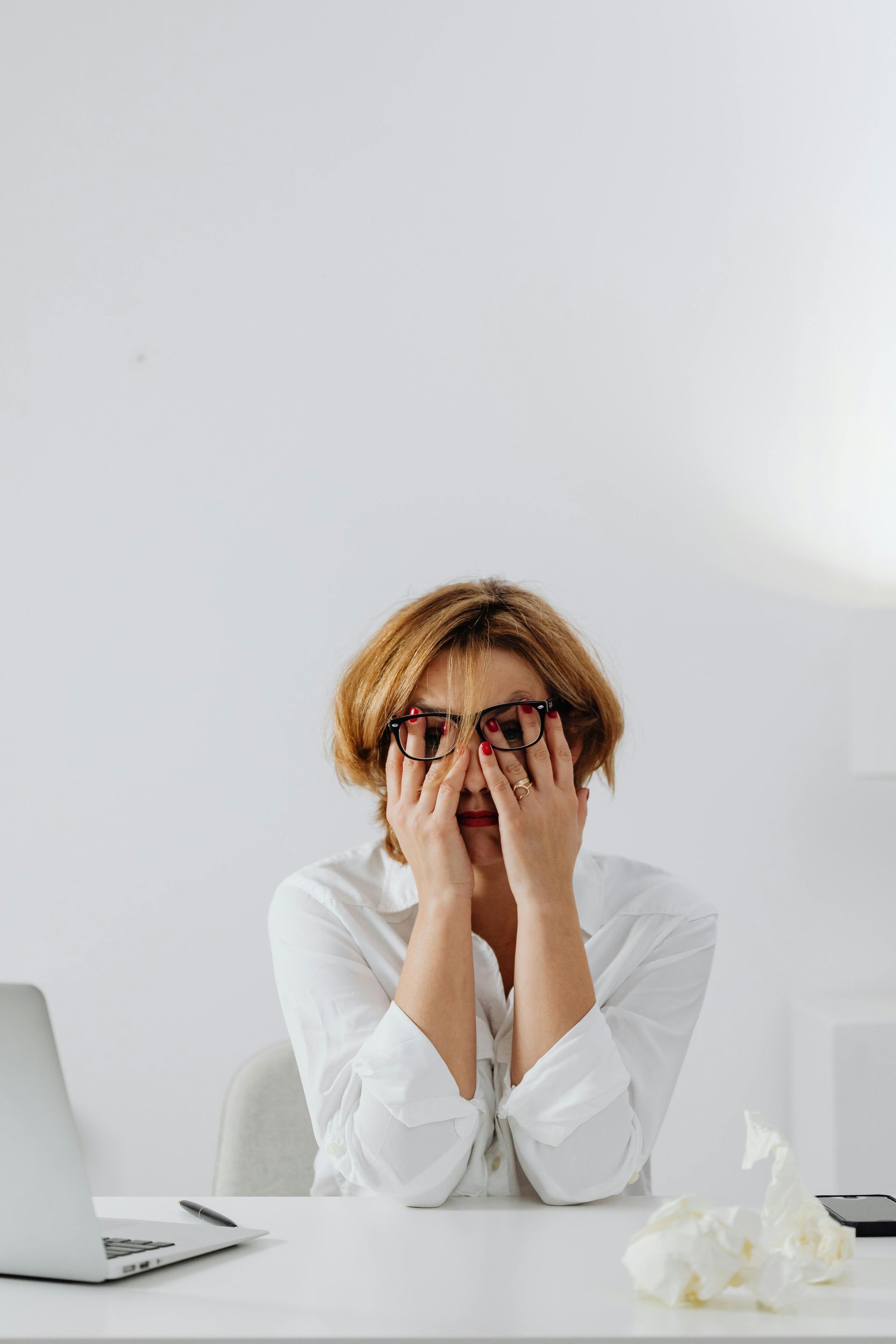 Woman covering eyes, feeling frustrated, in front of a white wall. 