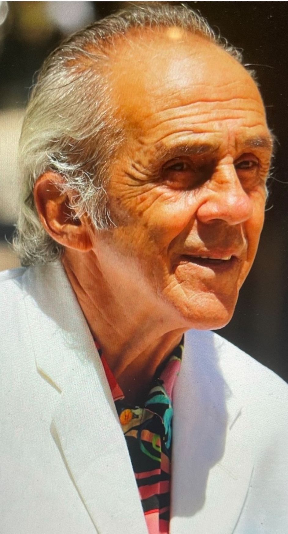 A man wearing a white jacket and a pink shirt is smiling.