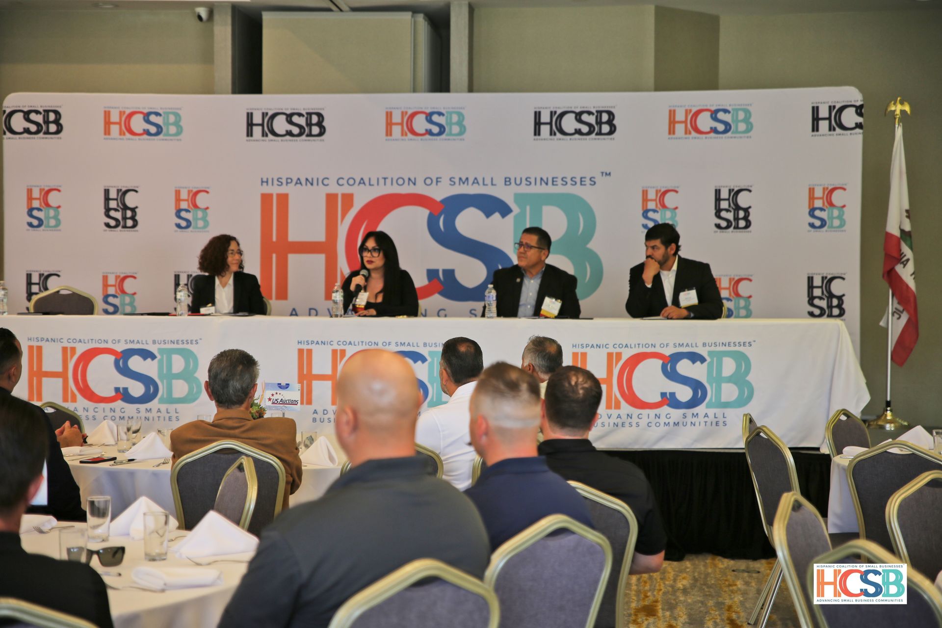 A group of people are sitting at tables in front of a sign that says hcsb