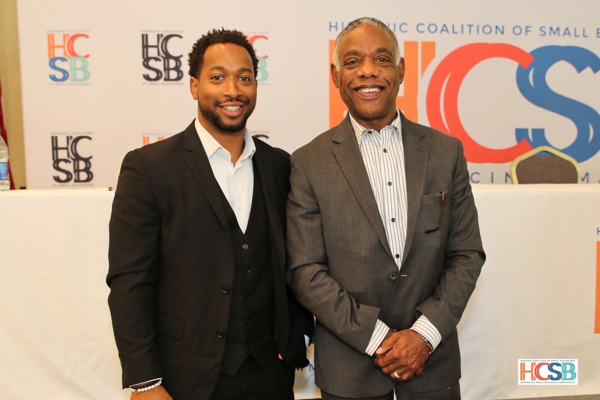 Two men are posing for a picture in front of a hcsb banner
