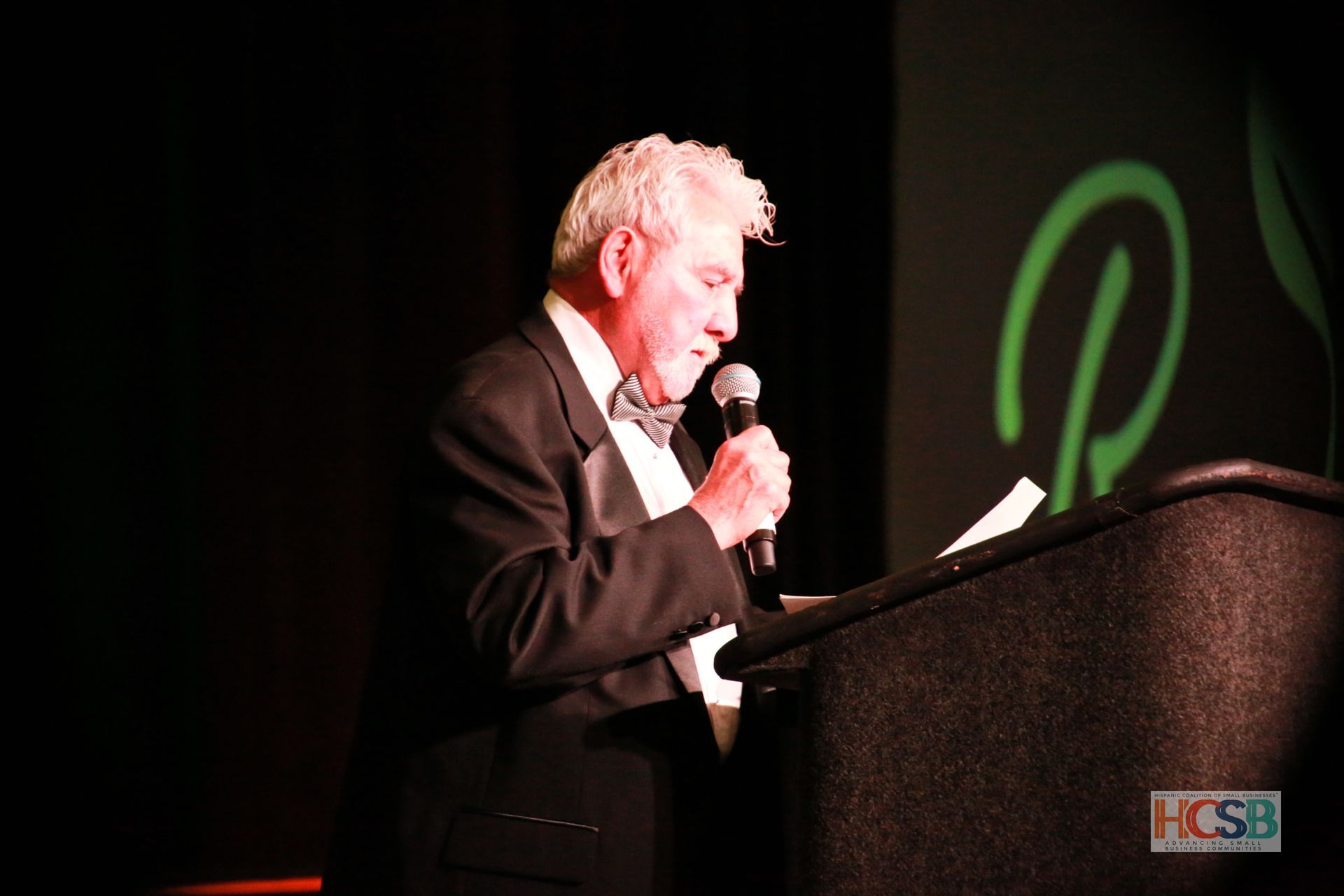 A man in a tuxedo is speaking into a microphone in front of a sign that says hcsb