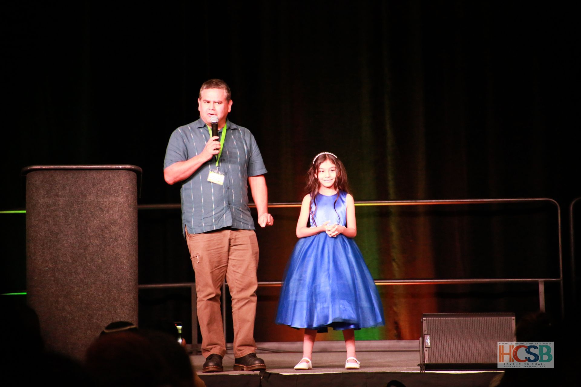 A little girl in a blue dress is standing next to a man holding a microphone on a stage.