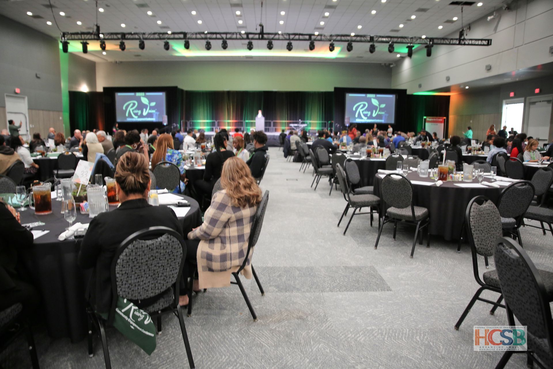 A group of people are sitting at tables in a large room with a hcsb logo in the corner