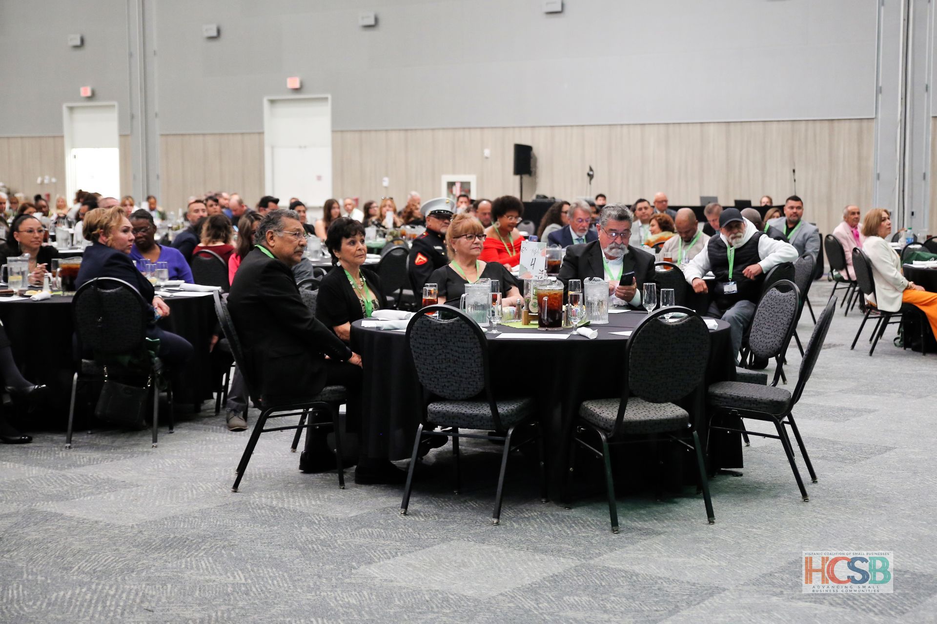 A large group of people are sitting at tables in a large room with hcsb written on the bottom