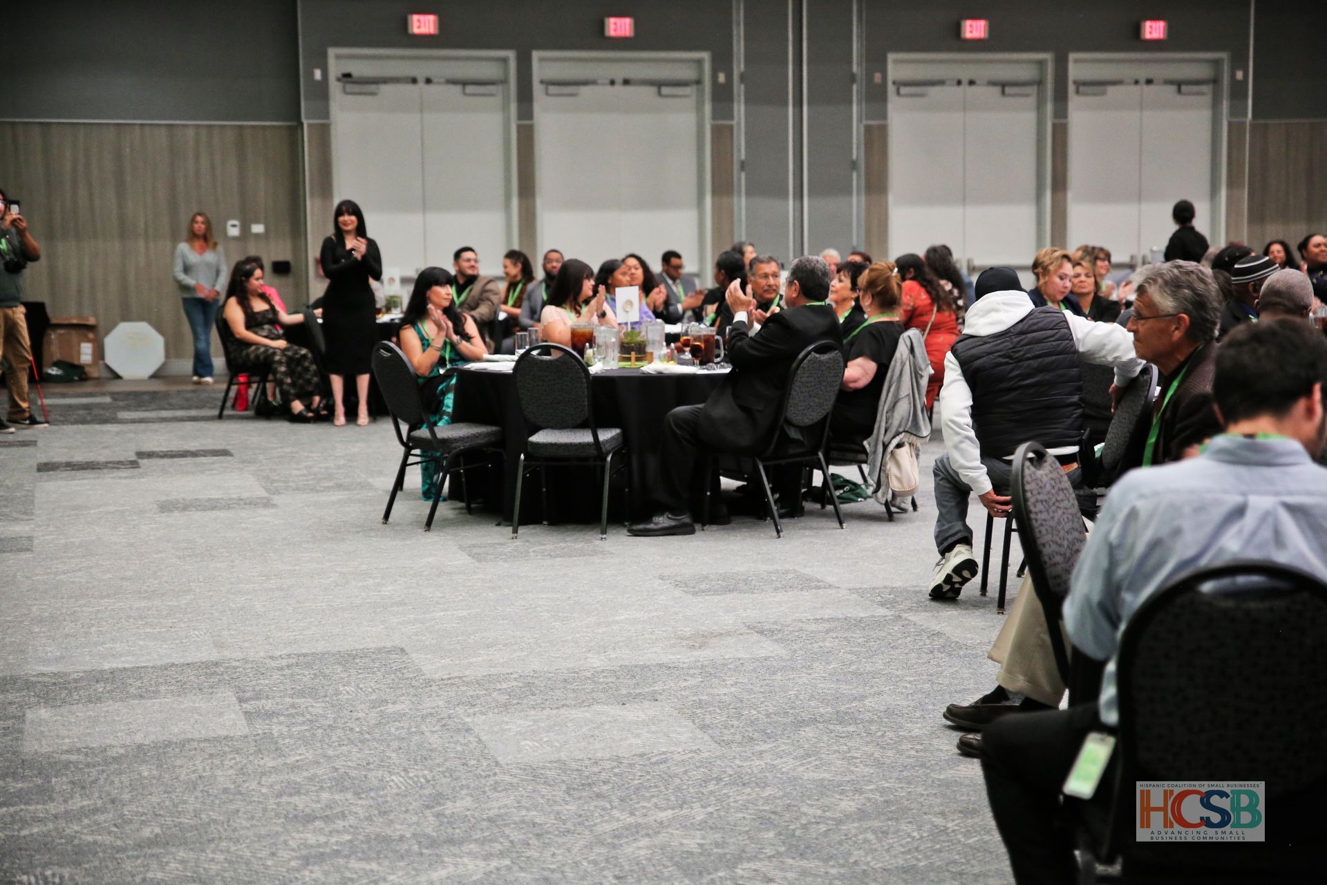 A group of people are sitting at tables in a room with a sign that says hcsb