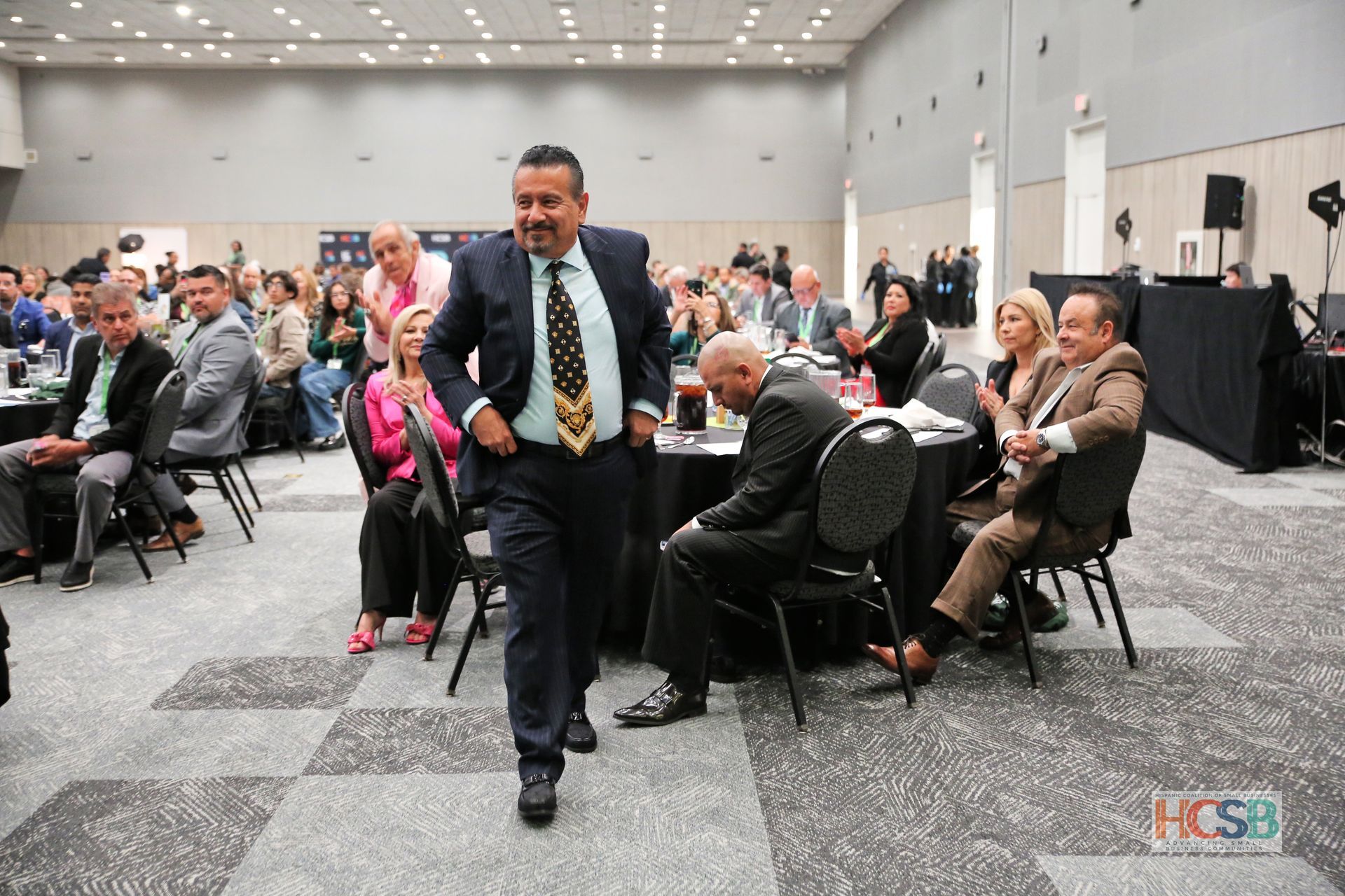 A man in a suit and tie is walking through a room with people sitting at tables.
