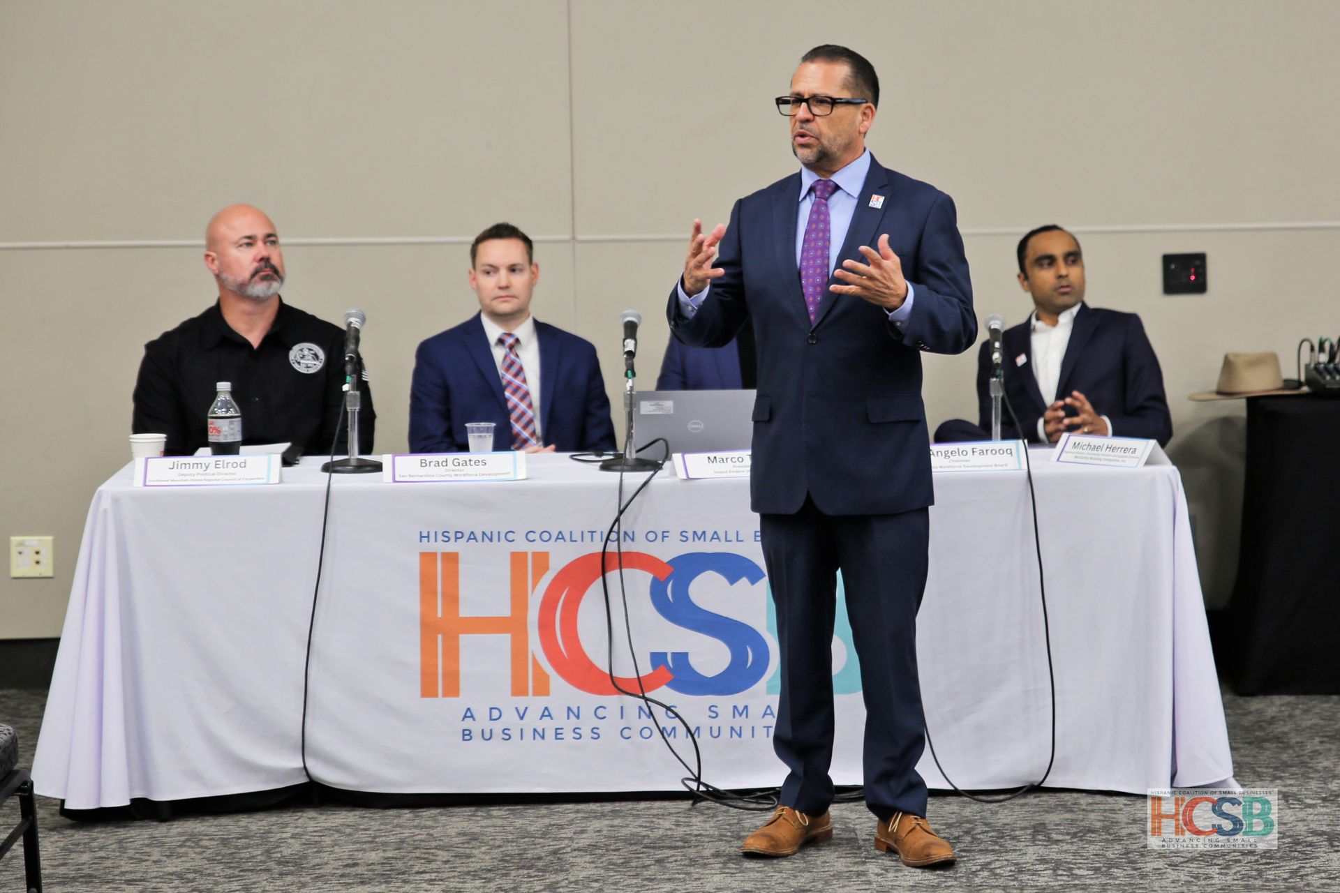 A man in a suit and tie is standing in front of a table with people sitting at it.