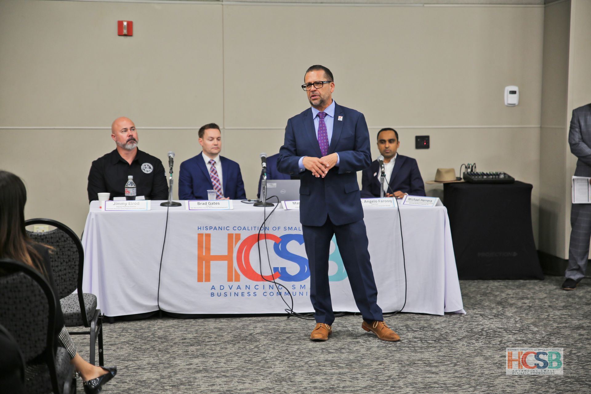 A man in a suit and tie is standing in front of a table with people sitting at it.