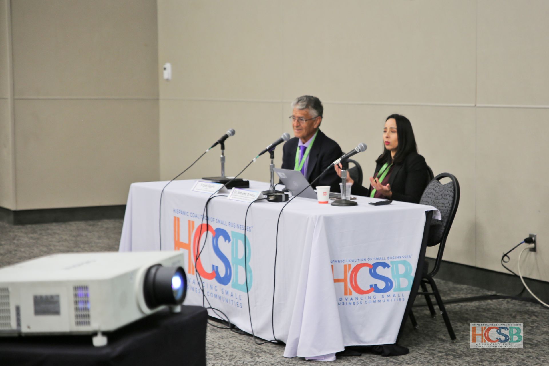 A man and a woman are sitting at a table with microphones and a projector that says hcsb