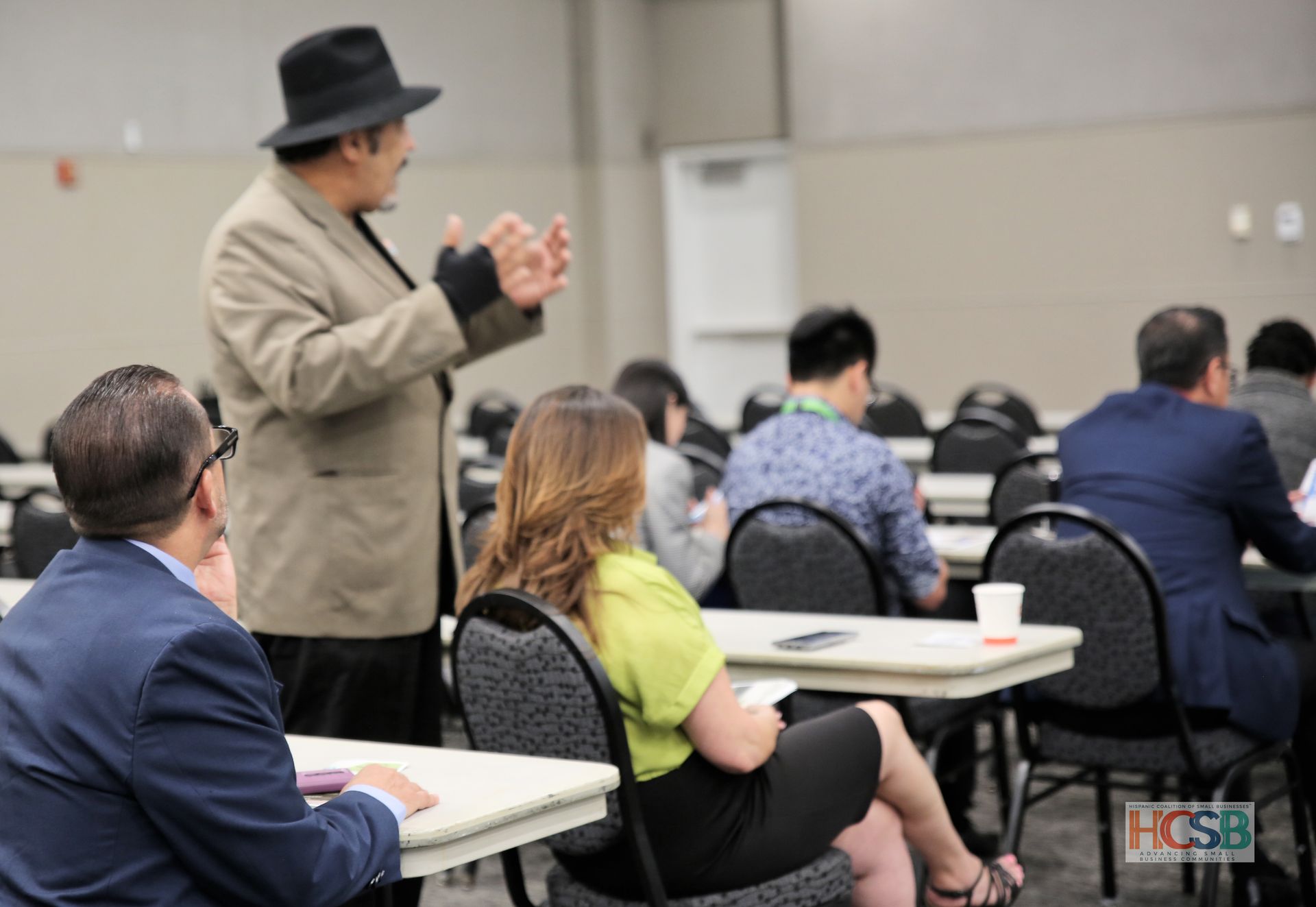 A man in a hat is talking to a group of people sitting at tables.