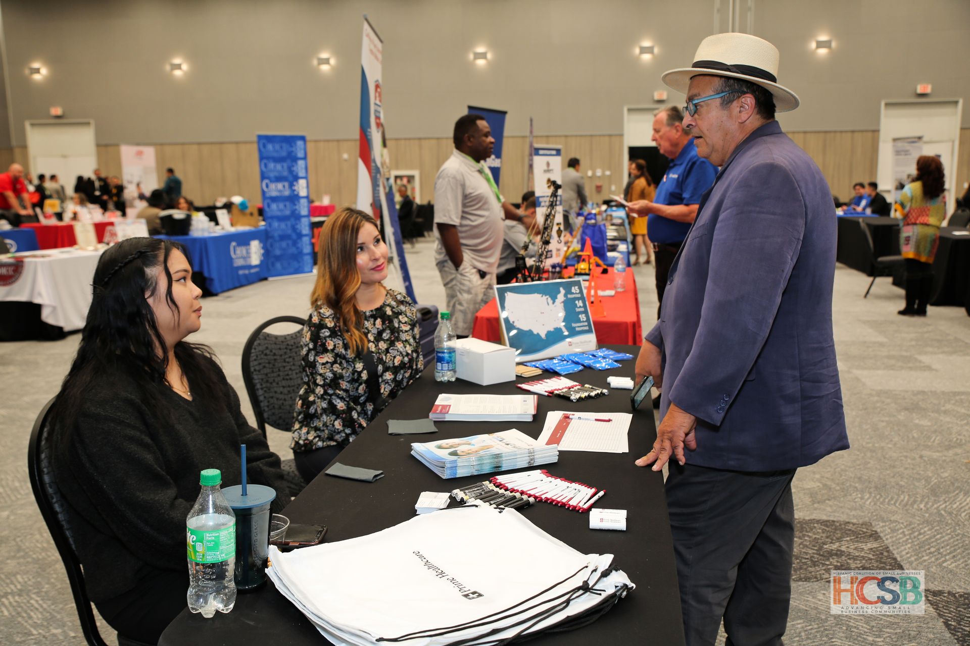 A man in a hat is talking to two women at a table at a job fair.