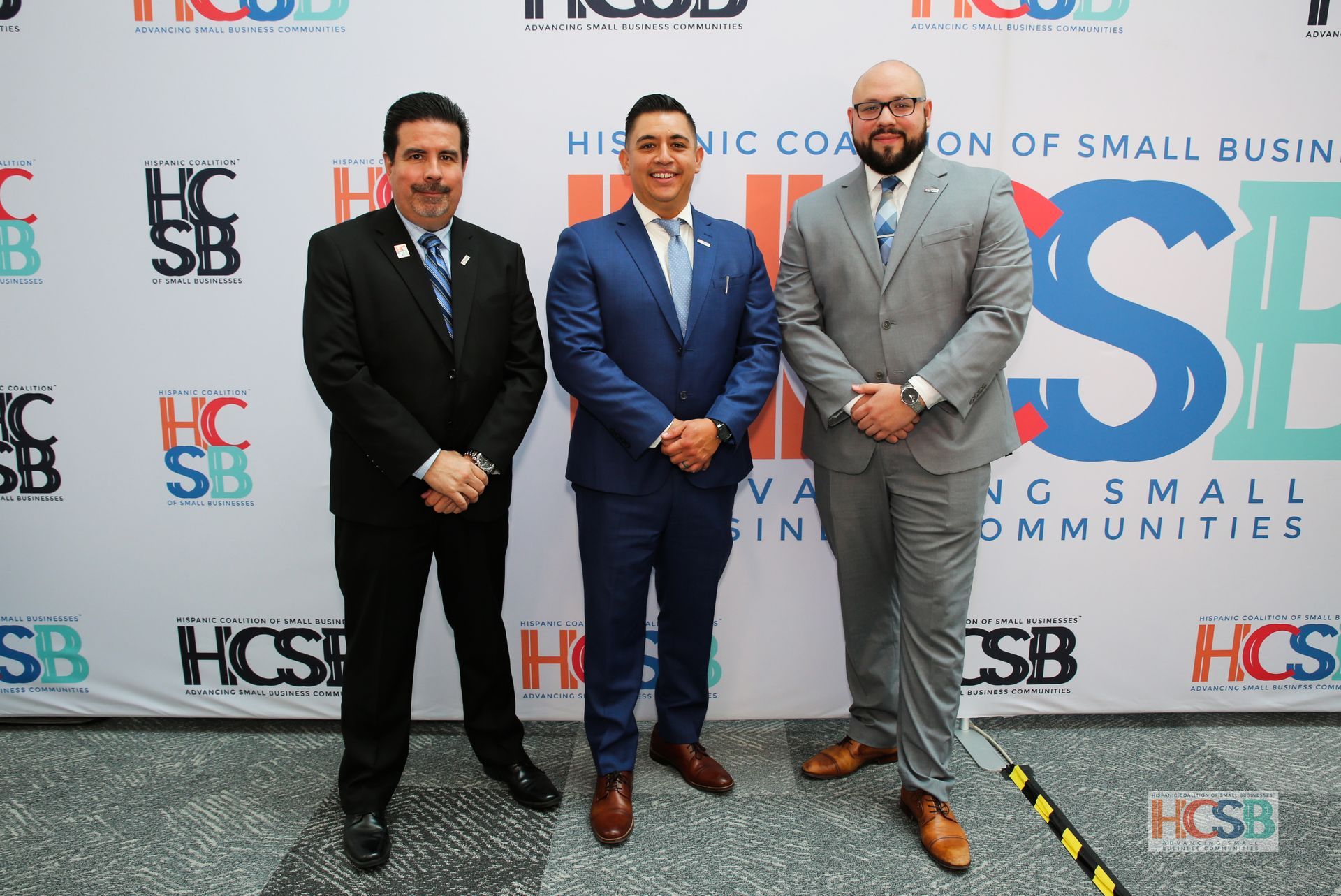 Three men in suits are posing for a picture in front of a wall that says hispanic coalition of small businesses.