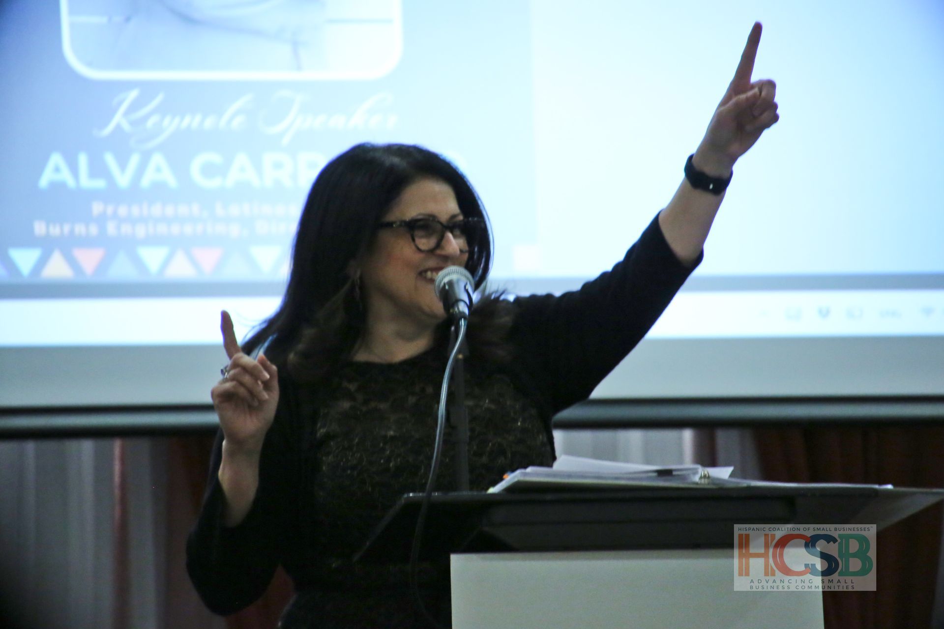 A woman is giving a speech at a podium with the name alva cary on the screen behind her