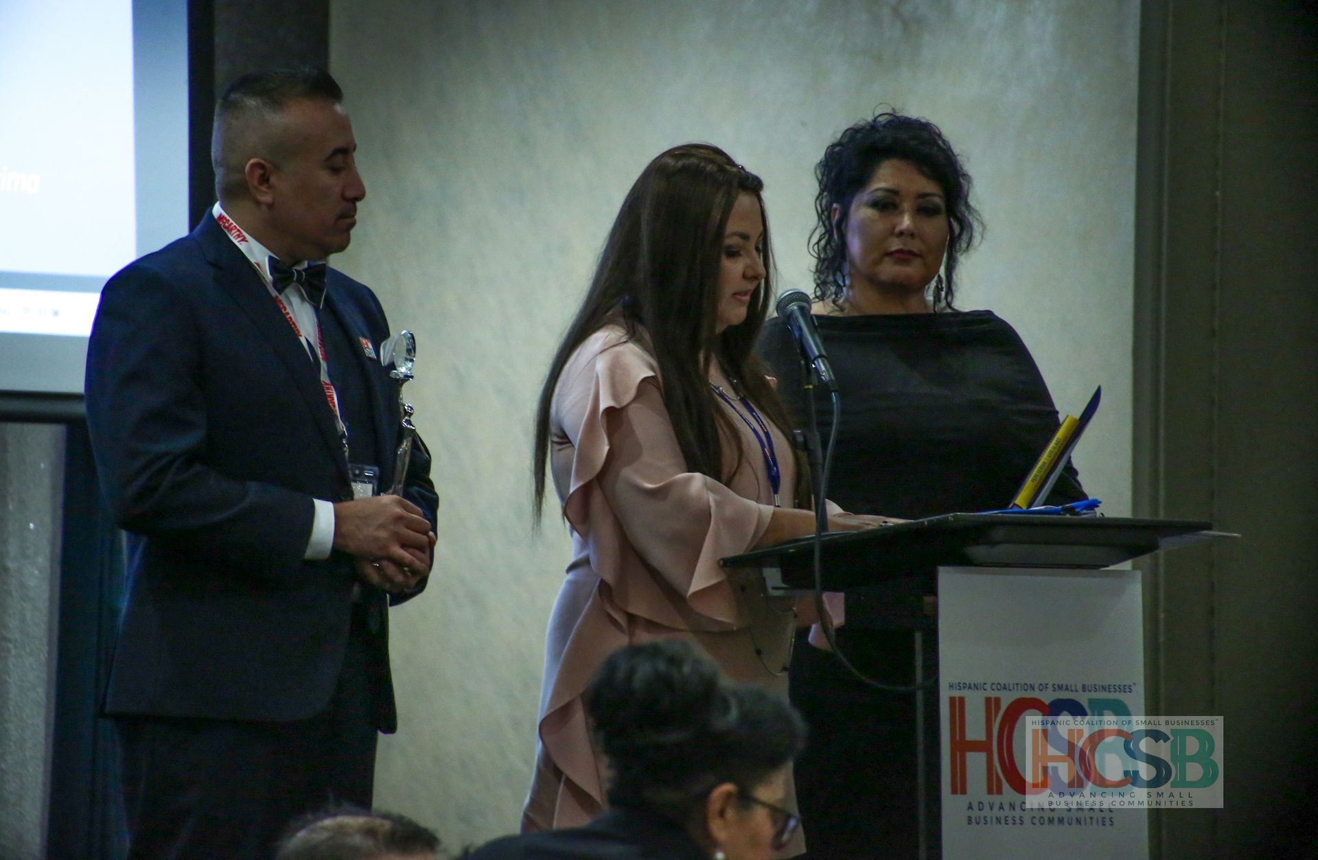 A woman is giving a speech at a podium while a man stands behind her.