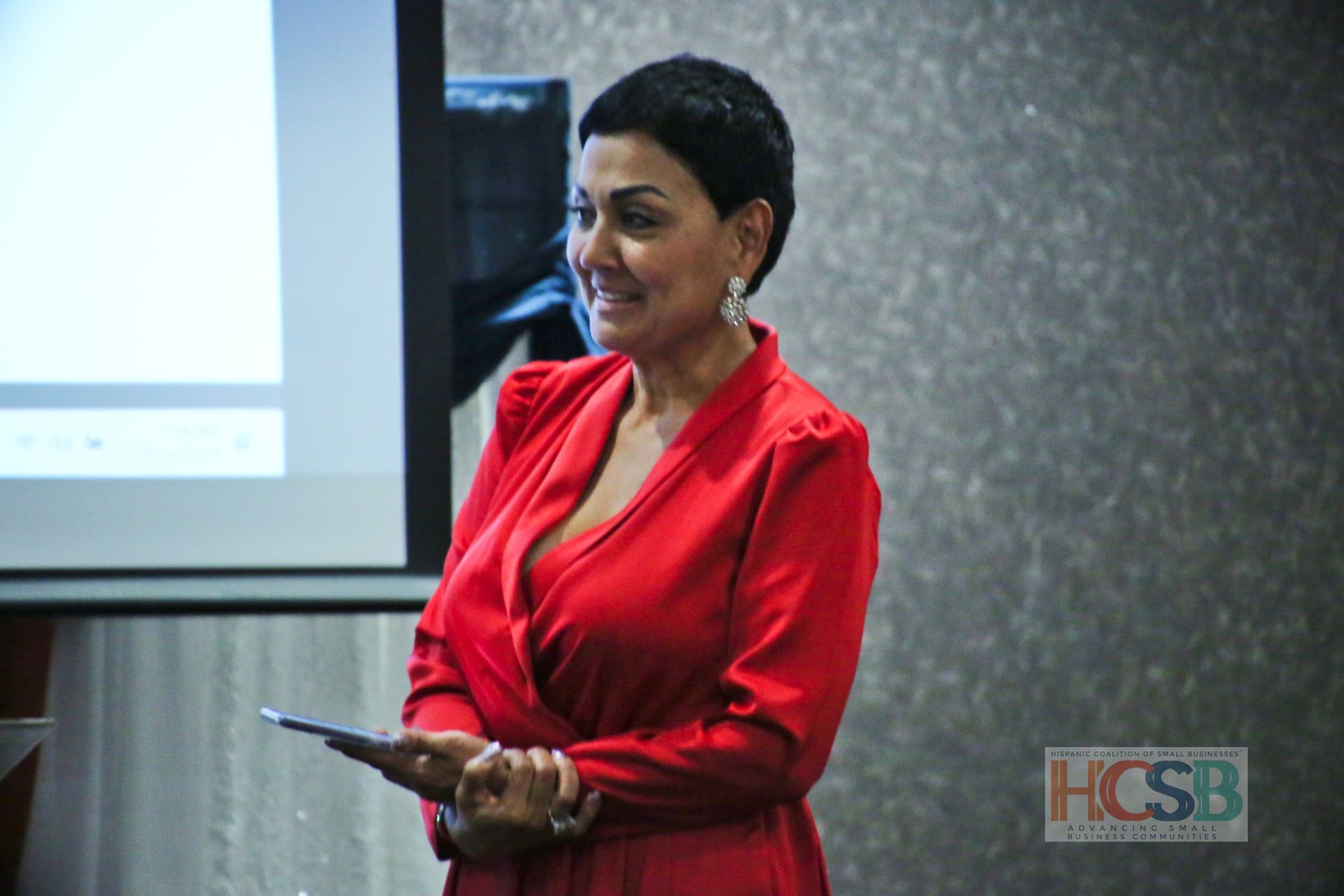 A woman in a red dress is standing in front of a projector screen holding a tablet.
