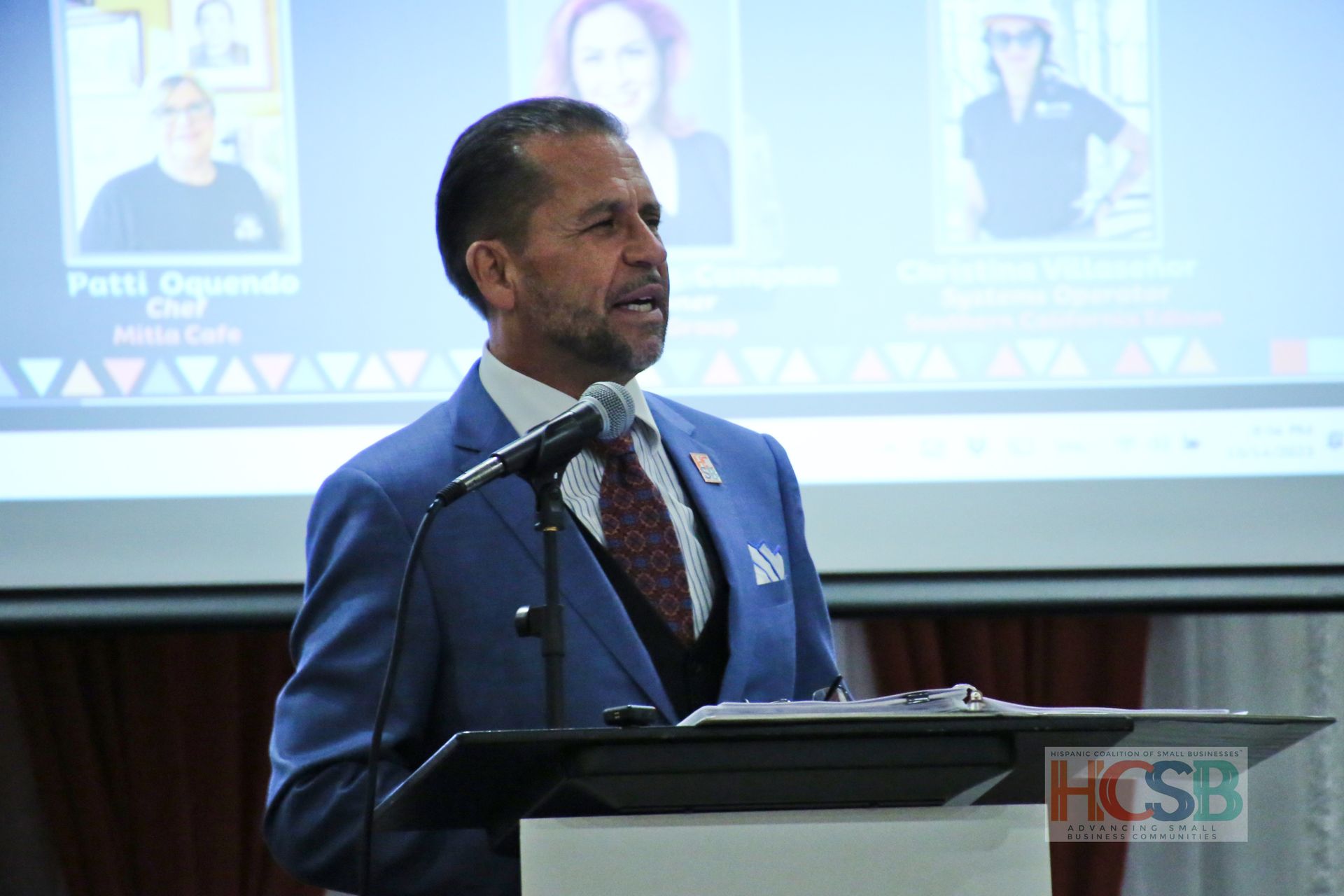 A man in a suit and tie is standing at a podium speaking into a microphone.