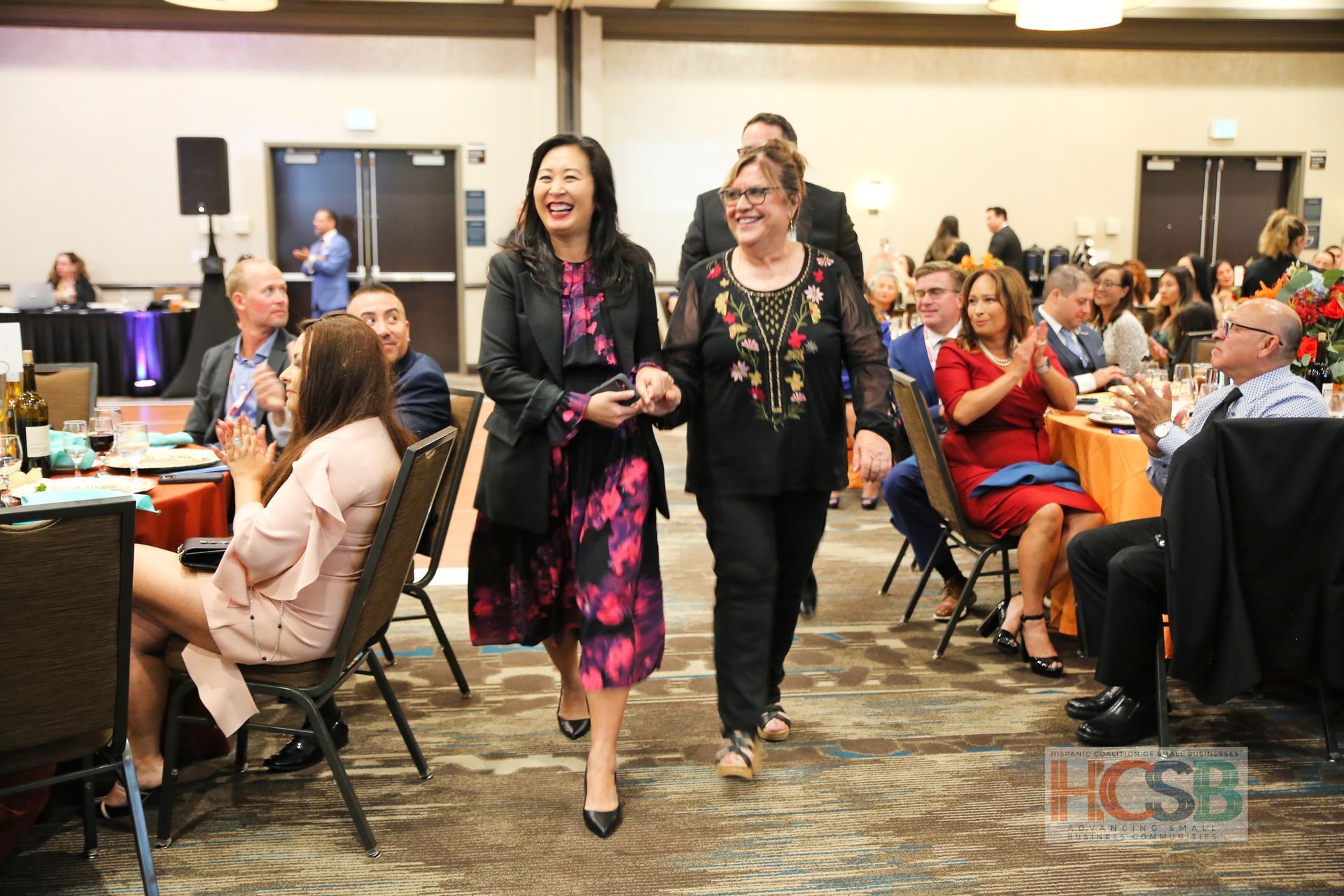 Two women are walking through a room with people sitting at tables.