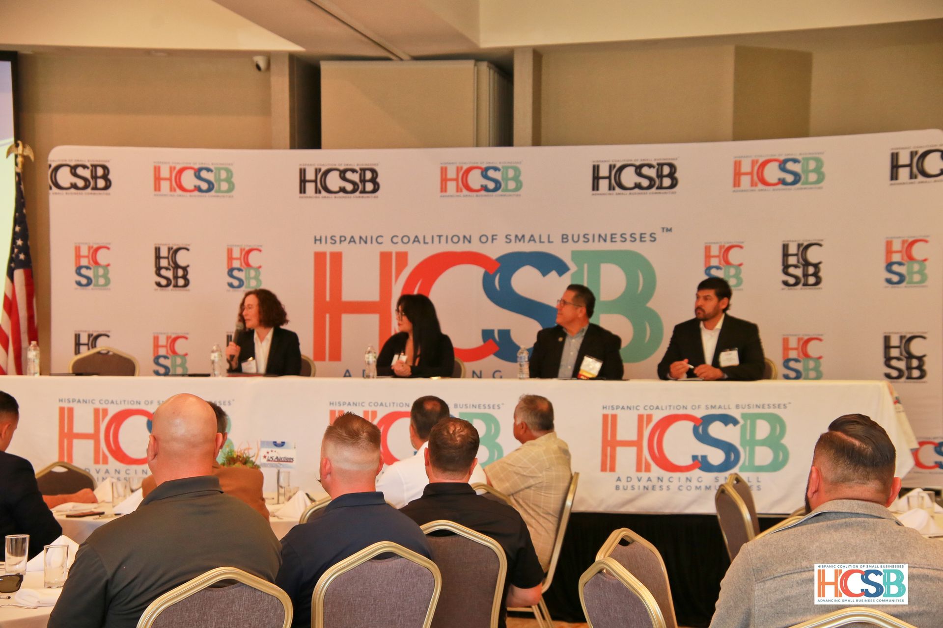 A group of people are sitting at tables in front of a wall that says hcsb.