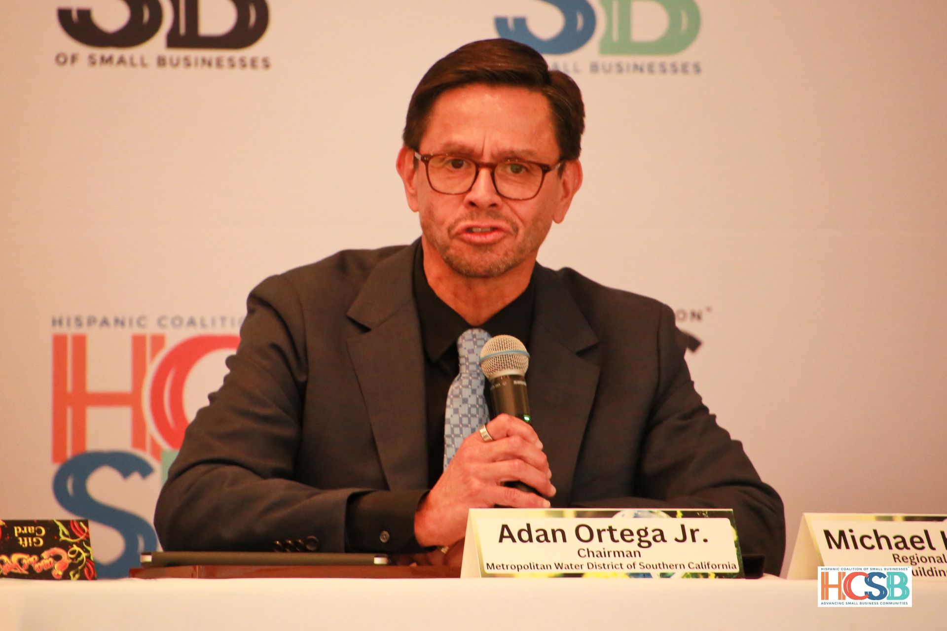 A man in a suit and tie is sitting at a table holding a microphone.