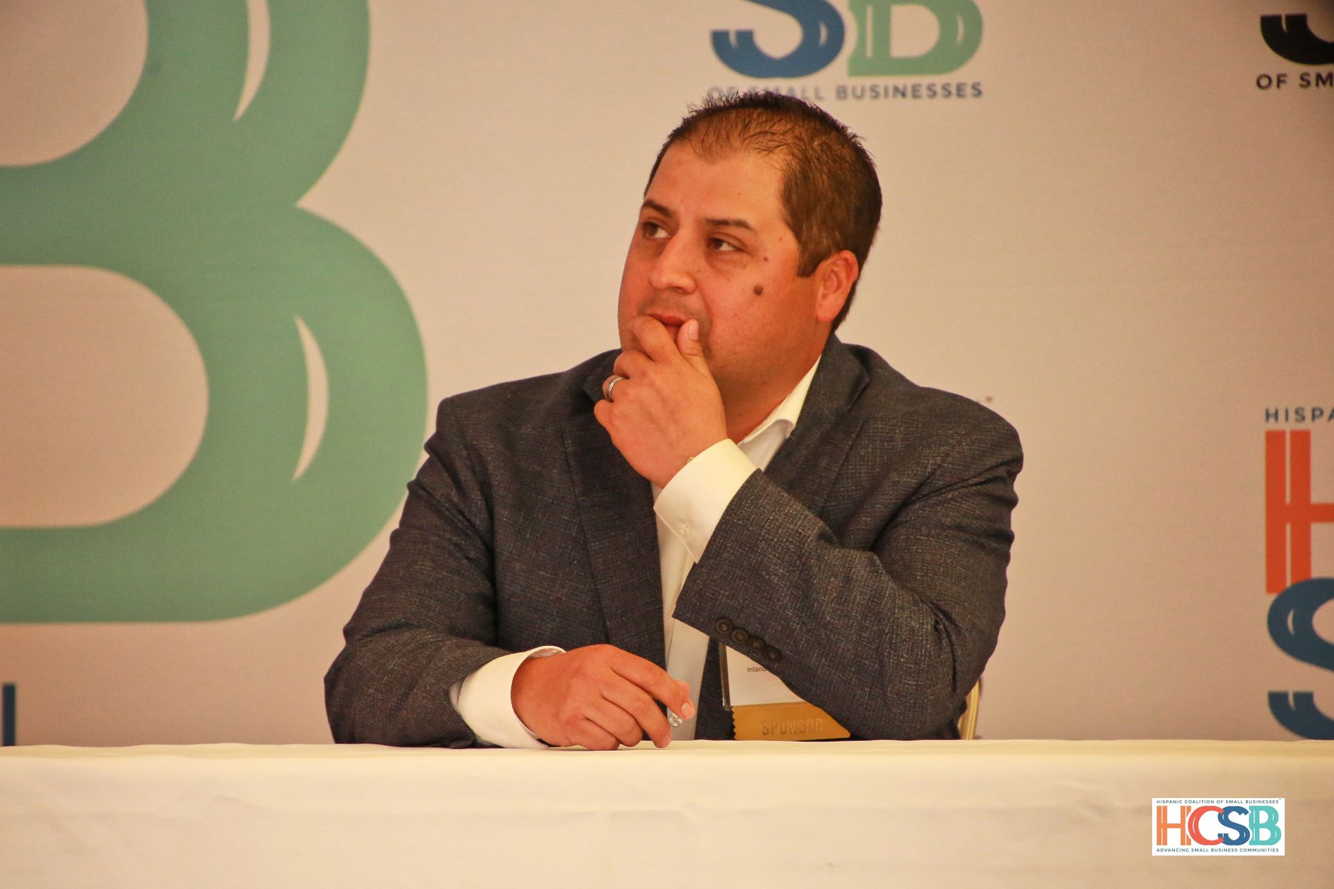 A man in a suit sits at a table with his hand on his chin
