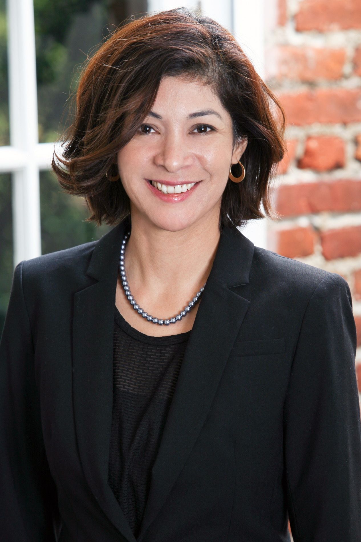 A woman in a black suit and pearl necklace is smiling in front of a brick wall.
