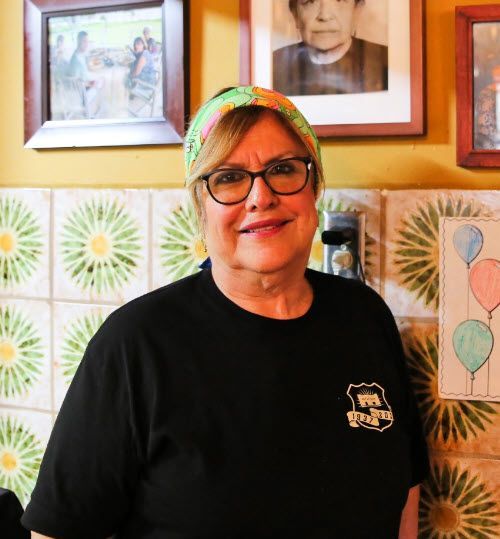 A woman wearing glasses stands in front of a wall with pictures on it