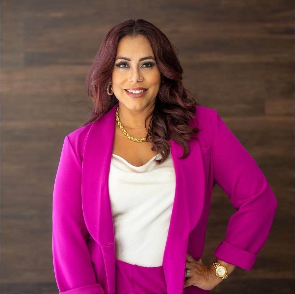 A woman in a pink jacket and white shirt is standing in front of a wooden wall.