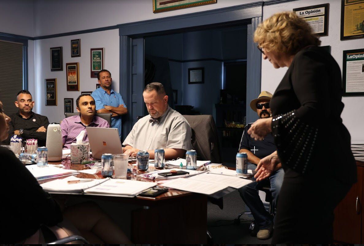 A woman is standing in front of a group of people sitting around a table.