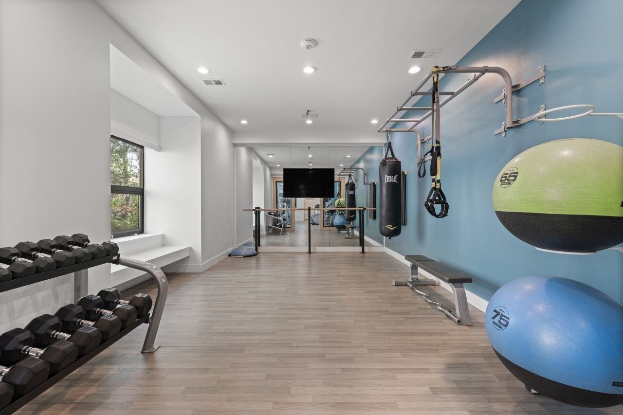 Well-lit apartment gym with dumbbells on racks, medicine balls, and a punching bag along a blue wall.
