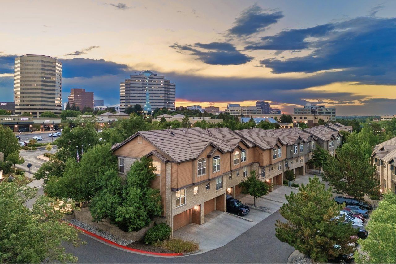 Exterior view of a townhouse-style apartment community with garages, trees, and a distant city skyline at sunset.