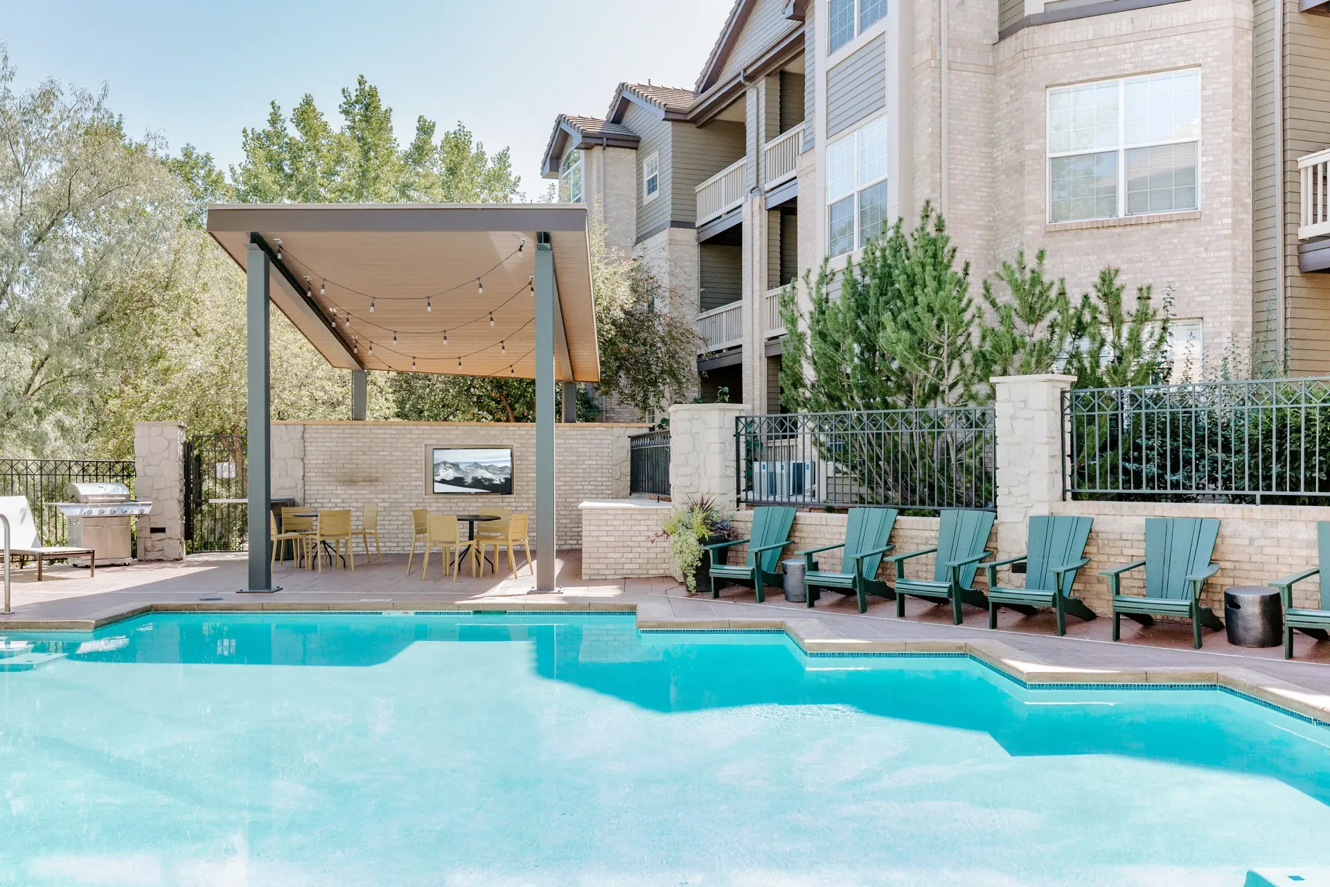 Outdoor communal pool with a covered pavilion, string lights, and teal lounge chairs near an apartment building.