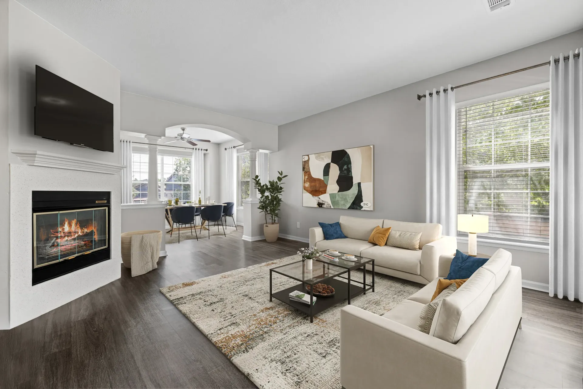 Modern apartment living room with a fireplace, wall-mounted TV, beige sofas, and a dining area by large windows.