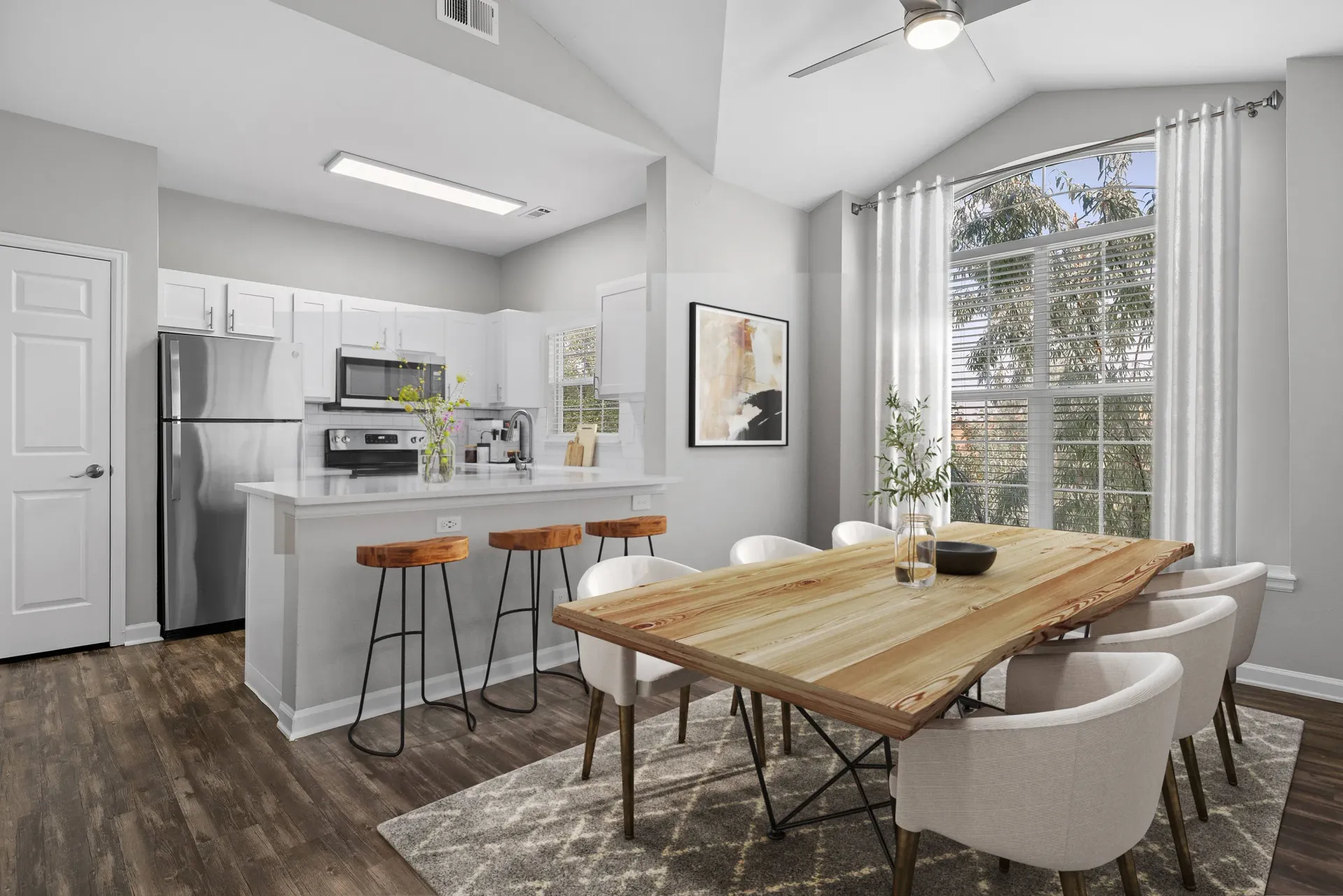Open-concept kitchen with island and bar stools, adjacent dining table, and a large window with white curtains.