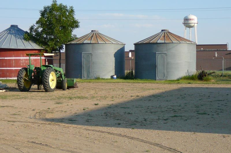 A green tractor is parked in a field with silos in the background